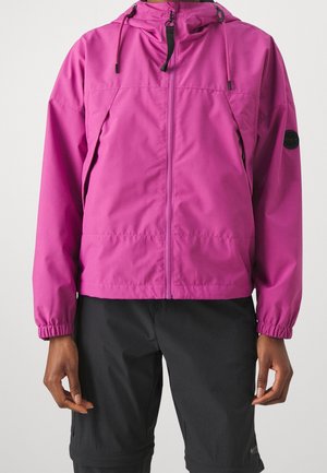 Osoba ubrana w jaskrawo różową bluzę z kapturem zapinaną na zamek, z elastycznymi mankietami, oraz czarne spodenki na jednolitym tle.