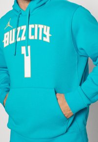 Sweat à capuche turquoise avec une poche devant, arborant "BUZZ CITY" et le numéro "1" en blanc et crème, avec un accent du logo Jordan.