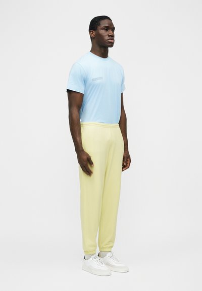 Mężczyzna stojący lekko zwrócony w prawo, ubrany w jasnoniebieską koszulę z krótkim rękawem, bladożółte spodnie dresowe i białe trampki na jednolitym białym tle.