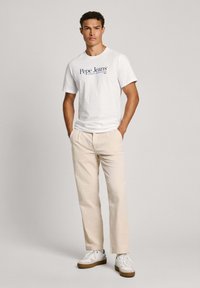 Balti trumpu rankovu marškinėliai su "Pepe Jeans" logo, derinami su šviesiais smėliniais kelnėmis ir baltomis sportinėmis avalynėmis su rudiu padu, stovinčioje pozoje.