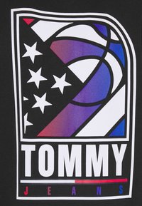 Logo a tema basket con stelle e strisce in sfumature di rosso, bianco, blu e viola, con il testo "TOMMY JEANS" sottostante.