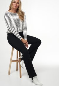 Zwarte jeans gecombineerd met een wit en marineblauwe gestreepte lange mouwen top. Het model draagt witte hoge hakken en zit op een houten kruk.