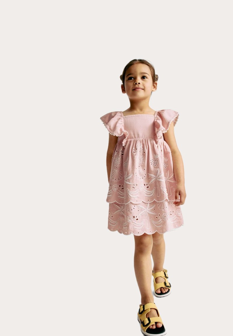 Robe en coton rose avec broderie à œillets, manches volantées et ourlet festonné. Associée à des sandales jaunes avec des accents noirs.