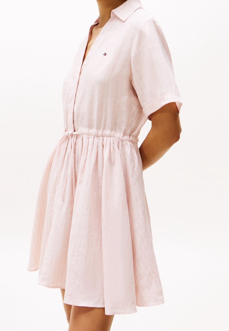 Femme portant une robe rose pâle à manches courtes avec col, boutons devant, taille cintrée et petit logo sur la poitrine, debout de profil.