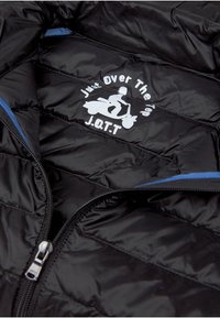 Giacca a puffo nera con dettagli blu, texture trapuntata e un logo bianco che raffigura una persona su uno scooter con la scritta "Just Over The Top J.O.T.T."