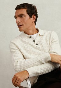 Kabelstrickpullover in Creme mit einem Kragen und drei schwarzen Knöpfen. Gerippte Bündchen und Saum verleihen dem Design Textur.
