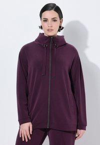 Sudadera con cremallera en color burdeos, con un cuello alto, detalles de cordón y bolsillos laterales. Está hecha de una tela suave con una textura lisa.
