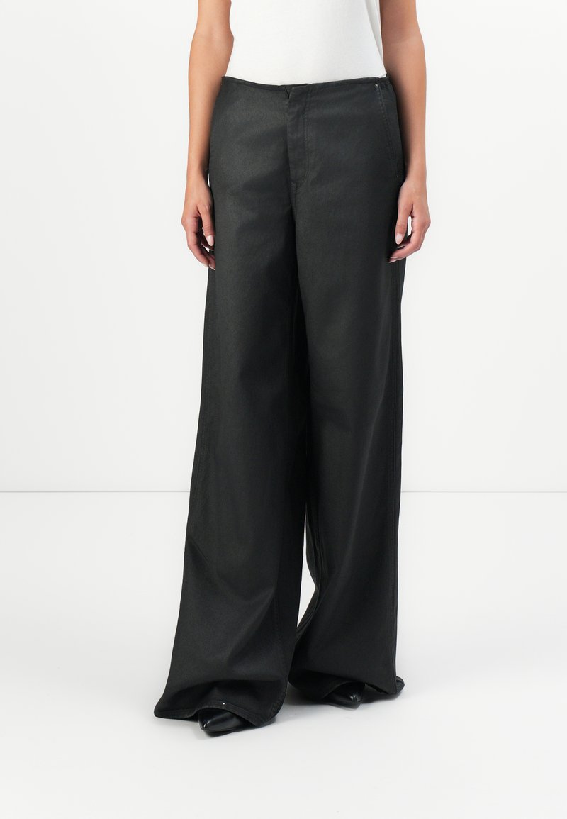 Personne portant un pantalon noir taille haute à jambes larges et des chaussures noires à bout pointu, se tenant devant un fond blanc uni.