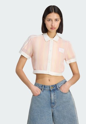 Kobieta o prostych ciemnych włosach, ubrana w różowy, prześwitujący, krótki top z białym kołnierzykiem i krótkimi rękawami oraz szerokie, wysokie niebieskie jeansy, z rękami w kieszeniach.