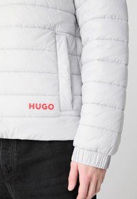 Gaišs pelēks pūkains džemperis ar kvadrātveida struktūru, elastīgām piedurknēm, sānu kabatu un izteiksmīgu sarkano "HUGO" logo tuvu apakšmalai.