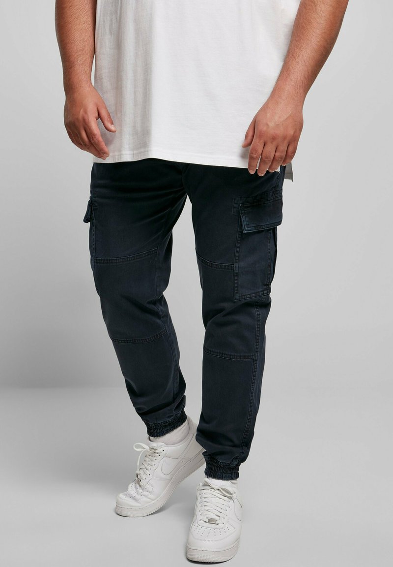 Mörkblå cargobyxor med flera sidofickor, avsmalnande anklar och elastiska muddar, ihop med vita sneakers och en enkel vit t-shirt.