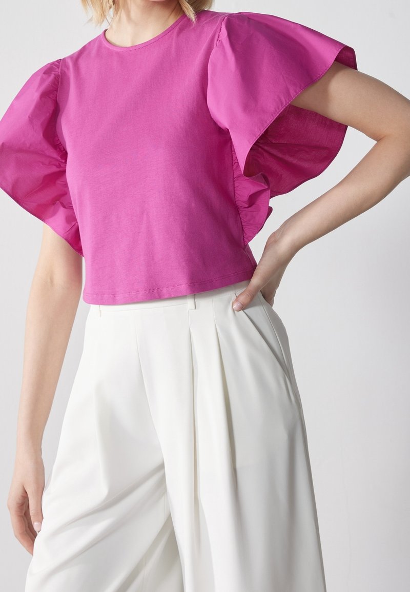 Femme portant un haut rose vif à manches courtes avec des manches volantées et un pantalon blanc taille haute à jambes larges, main sur la hanche.