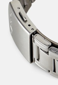 Metalen horlogeband met een geborstelde stalen afwerking, rechthoekige schakels en een sluiting met een drukknopmechanisme voor een veilige sluiting.