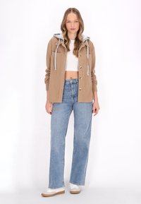 Jachetă din catifea călduță, maro, cu glugă gri și șnururi, cu închidere prin nasturi, asortată cu blugi din denim deschis, cu croială largă, și adidași albi.