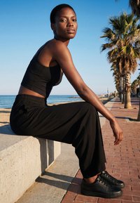 Woman in black crop top and wide pants sitting on concrete bench by beach promenade lined with palm trees under clear sky.