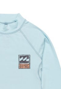 Rash guard de manga larga azul claro con cuello alto. Presenta un logo gráfico de olas en negro y naranja en el pecho y detalles de costura plana.