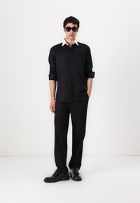 Svart skjorte med hvit krage, kombinert med svarte bukser og svarte sko; solbriller fullfører stilen. Enkelt, stilrent design.