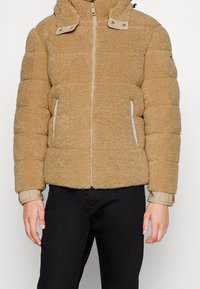 Tan faux shearling pufferjack met een hoge kraag, een ritssluiting aan de voorkant, twee zijzakken en elastische manchetten. Heeft een gestructureerd oppervlak.