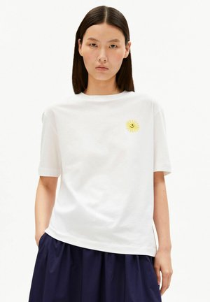 Woman with straight dark hair wearing a white t-shirt with a small yellow sun and black smiley face on the chest, paired with a dark blue skirt.