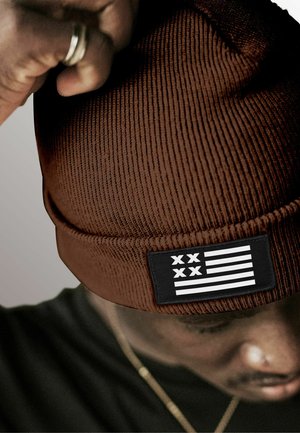 Brown ribbed beanie featuring a black patch with a white flag design, showcasing horizontal stripes and symbols. Textured and fitted style.