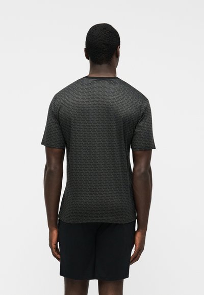 Homme vu de dos portant une chemise gris foncé à manches courtes avec un petit motif de texte noir et un short noir, debout devant un fond uni.