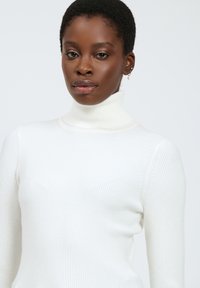 Maglione bianco a collo alto a costine con maniche lunghe, caratterizzato da una silhouette aderente e un tessuto testurizzato. Collo chiaro e liscio.