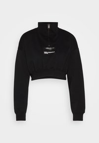 Svart kort tröja med hög krage, dragkedjestängning och en glansig märkespatch. Har ribbstickade muddar och en avslappnad passform.