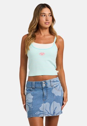 Jonge vrouw draagt een blauw-wit gestreept tanktopje en een blauwe denim minirok met grote bloemmotieven, staand tegen een effen achtergrond.