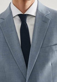 Veste de costume bleu clair avec un col cranté, assortie d'une chemise habillée blanche et d'une cravate bleu marine. Elle possède une poche poitrine unique.