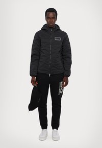 Sort quiltet jakke med hætte, lynlås og sidelommer; parret med sorte sweatpants med et hvidt logo og hvide sneakers.