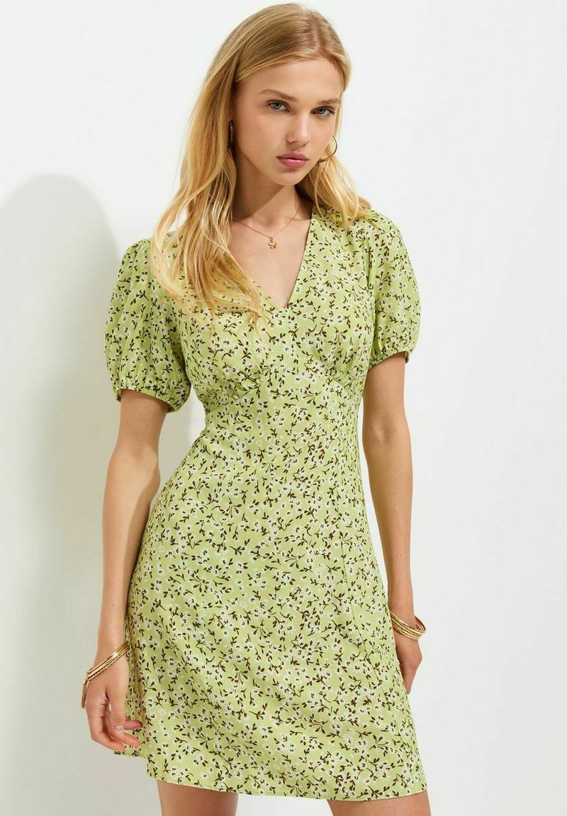 Woman with long blonde hair wearing a light green floral short-sleeve dress and gold bracelets standing against a plain white background.