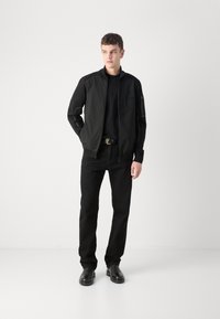 Black bomber jacket with zip front, ribbed cuffs, and a smooth texture. Worn over a black T-shirt and paired with black jeans and footwear.
