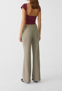 Vrouw in hoge taille, beige geruite wijde broek en een bordeauxrode top met open rug, staande tegen een effen lichte achtergrond.