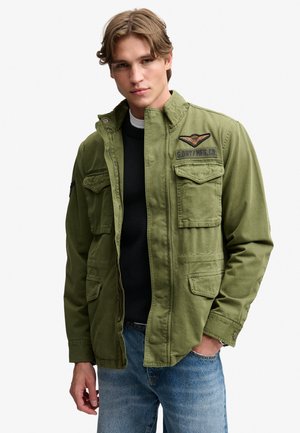 Ung mand iført en grøn militærinspireret jakke med flere lommer og mærker, der er unzipet, over en sort sweater og blå jeans.