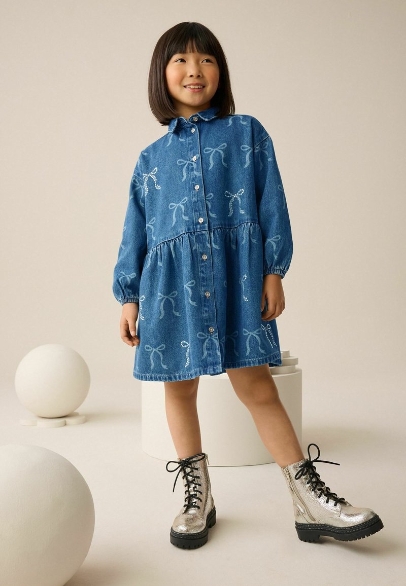 Denimkleid mit hellblauen Schleifenmustern, durchgehender Knopfleiste und elastischen Ärmeln. Kombiniert mit glänzenden silbernen Schnürstiefeln.