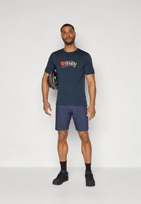 Tumši zils T-krekls ar logotipu, kopā ar tumšām šortām un melnām kedām. Modelis tur ķiveri, stāvot pret vienkāršu baltu fonu.