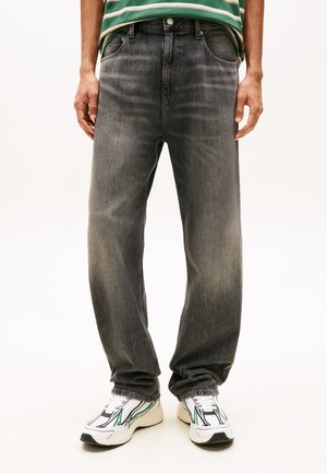 Személy kopott fekete bőszárú farmerben, zöld-fehér csíkos ingben és zöld-fekete részletekkel ellátott fehér tornacipőben.