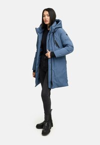 Donna che indossa un cappotto invernale blu con cappuccio, maglione nero a collo alto, jeans neri e stivali neri spessi, in piedi e rivolta in avanti.