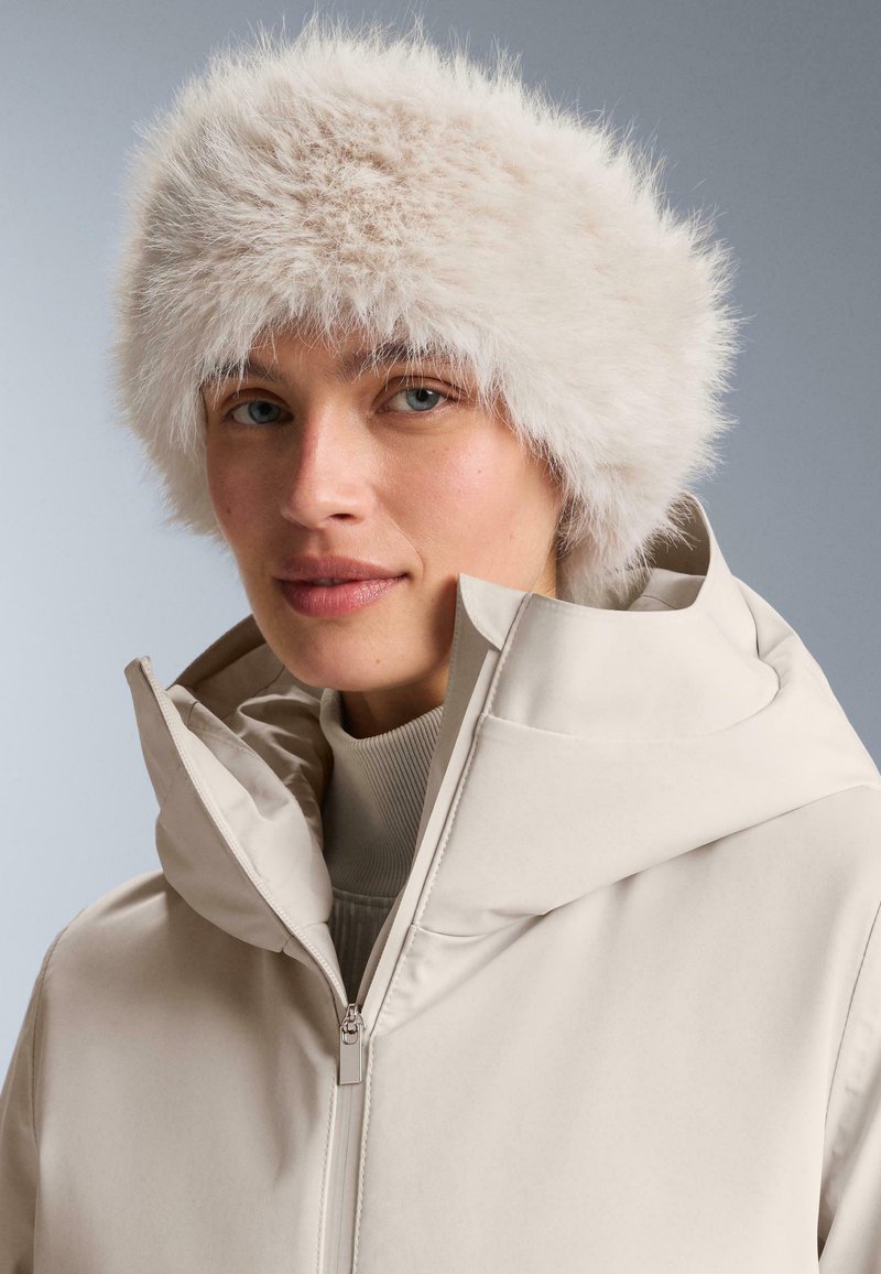 Fehér műszőrme sapka lekerekített formával, világos bézs cipzáras dzsekivel viselve, sima textúrával és modern sziluettel.