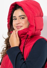 Rote, gepolsterte Weste mit hohem Kragen und verstellbarem Kapuze, ausgestattet mit schwarzen Kordeln. Darunter ist ein marineblaues Langarmshirt sichtbar.