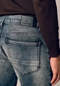 Spodnie dżinsowe w kolorze wyblakłej szarości o fakturowanej powierzchni, z tylną kieszenią i czarną skórzaną metką z napisem "HENSEN" przy pasku.