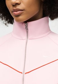 Κοντινό πλάνο ενός ατόμου που φοράει ένα ανοιχτό ροζ ζακέτα με φερμουάρ και ψηλό γιακά, με μια κόκκινη διαγώνια ρίγα στο στήθος.