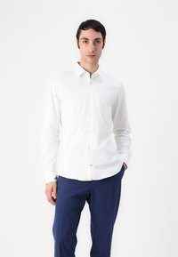 Chemise blanche à manches longues avec col boutonné, en tissu lisse, associée à un pantalon bleu foncé. Design simple, sans motifs visibles.