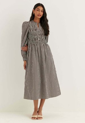 Donna che indossa un abito midi vichy bianco e nero con maniche lunghe a sbuffo e vita arricciata, abbinato a sandali beige con punta aperta.