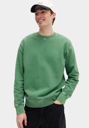 Lachende man met een lichtbeige pet, groene rondehals trui en zwarte broek, met één hand in de zak tegen een effen achtergrond.