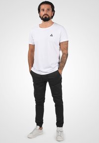 White cotton T-shirt with a round neckline and small black triangle logo, paired with black tapered pants and white sneakers.