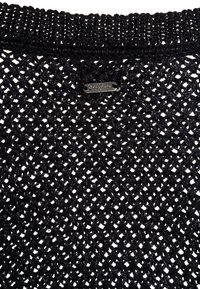 Černý pletený top se vzorem otevřeného tkaní. Okraje jsou žebrované a poblíž výstřihu je připevněná malá metalická značka.