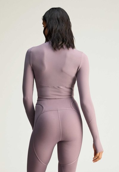 Woman with shoulder-length dark hair wearing a fitted mauve long-sleeve top and high-waisted leggings, viewed from the back.