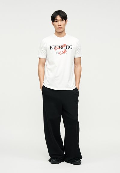 Νέος άνδρας στέκεται με τα χέρια στις τσέπες, φορώντας λευκό γραφικό μπλουζάκι με την επιγραφή «ICEBERG» και φαρδιά μαύρα παντελόνια, μπροστά σε απλό φόντο.
