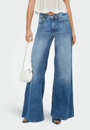 Kobieta w wysokich, szerokich niebieskich dżinsach i białym szydełkowanym topie na krótko, niosąca małą białą torebkę.